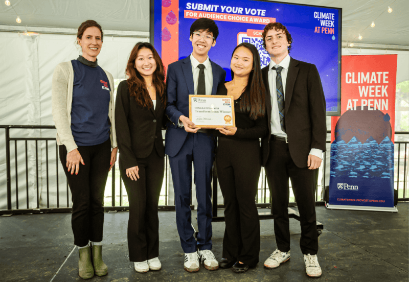 The image shows students on stage with a Climate Week banner behind them. The student in the middle holds a plaque.